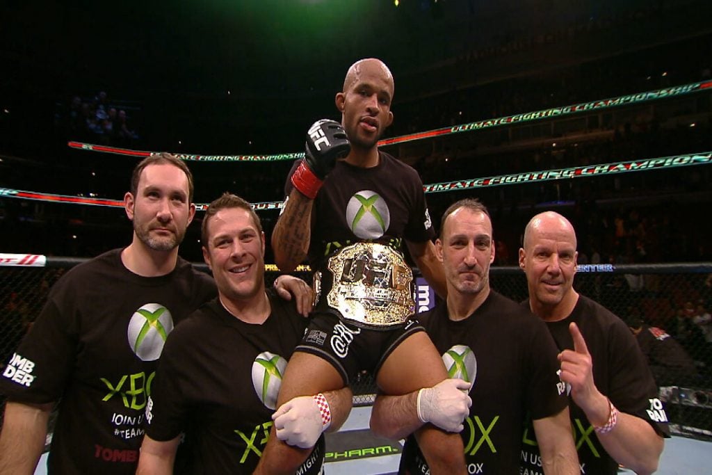 Dois homens erguendo o melhor lutador do UFC na história dos pesos-mosca, Demetrious Johnson.